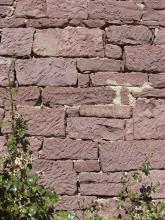 Blick auf eine aus rötlich grauen Steinen errichtete Mauer. Die Steine sind unterschiedlich groß und haben schräg verlaufende Furchen.