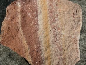 Handstück eines Bruchsteines mit senkrecht verlaufenden Farbstreifen von hellrot, dunkelrot, gelb und rosa.