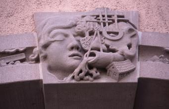 Moderne, aus rötlich grauem Stein gefertigte Reliefarbeit an einer Hauswand. Sie zeigt unter anderem ein Gesicht, einen Zirkel, ein Wasserrohr und rechts unten ein kleines Haus.
