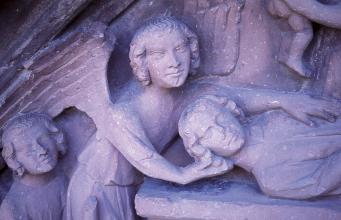 Nahaufnahme einer aus rötlich grauem Stein gehauenen Figurengruppe. Die mittlere von drei Figuren trägt Engelsflügel auf dem Rücken.