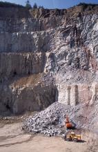 Blick auf eine sehr hohe, über mehrere Stufen gehende Steinbruchwand aus grauem bis rötlich braunem Gestein. Rechts unten arbeiten Bagger und Radlader vor einer Abraumhalde.