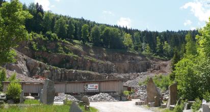 Blick auf ein Steinbruchgelände mit breiten, mittelhohen Gesteinsterrassen und dicht bewaldeter Kuppe. Im Vordergrund Lagergebäude und Gesteinshaufen sowie als Schaustücke aufgestellte Steinblöcke.