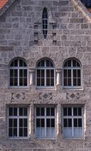 Ansicht einer hohen Hausfassade aus rötlich grauem Mauerwerk mit zwei Fensterreihen, einem Giebelfenster und kleineren Verzierungen.