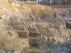 Blick auf eine hohe Steinbruchwand aus hellbraunem Gestein. Im unteren Teil sind Blöcke herausgearbeitet. Die oberen Wände sind links gelblich, rechts bläulich grau und darüber rotbraun. Auf der Kuppe steht Wald.