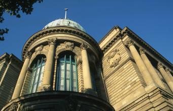 Teilansicht vom obersten Stockwerk eines Gebäudes mit gelblich braunem Mauerwerk. Im Bild ist ein runder, turmartiger Erker sowie rechts ein eckiger Anbau mit Säulen um die Fenster.  
