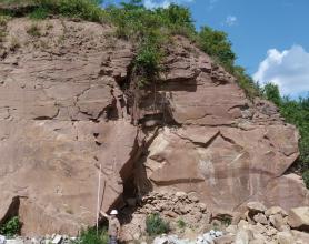 Blick auf eine mächtige, rötlich graue Steinbruchwand mit senkrechtem, gezacktem Riss in der Mitte und bewachsener, nach rechts abfallender Kuppe. Am unteren Bildrand hält ein Arbeiter eine Messlatte an das Gestein.