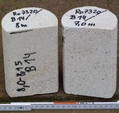 Nahaufnahme zweier kurzer runder Steinkörper, beide hellgrau mit kleinen bräunlichen Punkten. Beide Gesteine sind an der Frontseite gerade abgeschnitten sowie auf der Oberseite (links zusätzlich auf der Frontseite) beschriftet. Unten liegt eine Messlatte.