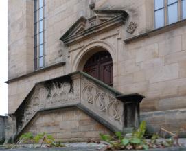 Blick auf ein Kirchenportal aus hellbraunem Mauerwerk, mit seitlichem Treppenaufgang und verziertem, steinernen Geländer.