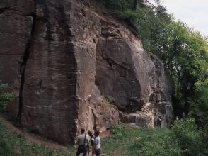 Blick auf eine massige, dunkle Felswand, die sich vor einem Waldhang befindet. Links durchzieht eine senkrechte Kluft das Gestein, rechts der Bildmitte ist eine größere Nische im Fels. Zwei Personen stehen am Fuß der Felsformation.