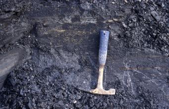 Das Bild zeigt ein dunkelgraues bis schwarzes Gestein. Im Vordergrund mittig vom Bild ist ein Geologenhammer als Maßstab plaziert.