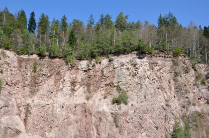 Steinbruch im Granit bei Alpirsbach – Auflagerung Deckgebirge über Grundgebirge Blick auf eine rötlich graue, hohe und lange Abbauwand in einem Steinbruch. Die wellige Kuppe ist bewaldet.