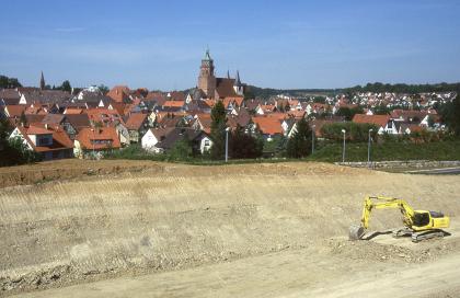 Abgrabung in Mergelsteinen des Unteren Muschelkalks bei Weil der Stadt Im Vordergrund des Bildes ist ein großer, gelblich brauner und grauer Hügel zu sehen, den ein Bagger an Sohle und Seite bearbeitet hat. Direkt dahinter breitet sich eine Stadt aus.