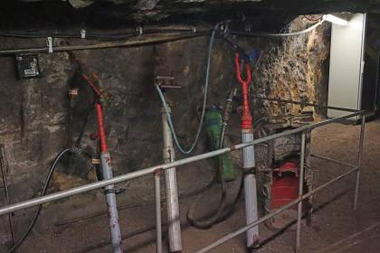 Druckluftbohrmaschinen im Silberbergwerk Teufelsgrund bei Münstertal Das Bild zeigt mehrere historische Bohrmaschinen im Besucherbergwerk Teufelsgrund. Die Bohrgeräte sind mit Luftschläuchen verbunden und stehen hinter einem Schutzgitter an einer Stollenwand.