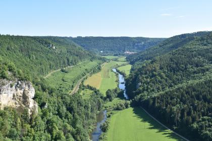 Das Obere Donautal bei Beuron – Blick vom Knopfmacherfels Blick aus großer Höhe über ein langgezogenes Tal mit Flusslauf. Das Tal ist hufeisenförmig von bewaldeten Bergen eingerahmt. Am Berghang links im Vordergrund stehen bleiche Felsen heraus.