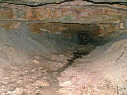 Blick in den Silberstollen südlich von Spiegelberg-Neulautern Das Foto zeigt das Innere eines Stollens. Zu sehen sind wannenförmige, graue Wände, eine rötlich braune, tiefhängende Decke sowie das trichterförmige Ende des Stollens.