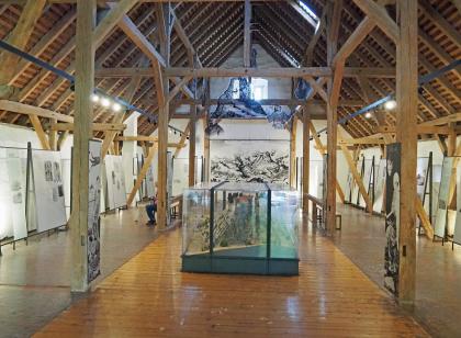 Ausstellung im Infozentrum zur Festungsruine Hohentwiel Das Bild zeigt einen Ausstellungsraum mit hoher, offener Balkendecke. In der Mitte ist ein Geländemodell sowie Zeichnungen des Hohentwiel zu erkennen. Links und rechts, in Seitengängen, sind Bildtafeln aufgestellt.