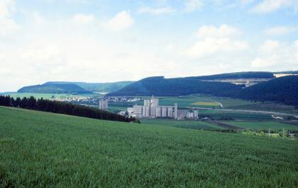 Donautal bei Geisingen von Südosten Zwischen einem nach rechts abfallenden grünen Acker und im Hintergrund wieder ansteigenden bewaldeten Höhen liegt eine grüne Mischlandschaft mit Siedlung und Werksgebäuden im Mittelgrund. Rechts oben ragt aus dem Wald ein Steinbruch hervor.