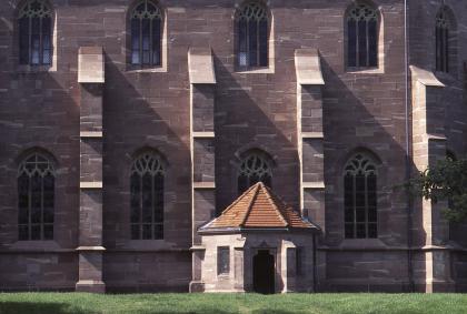 Kloster Hirsau bei Calw Das Bild zeigt die rötlich graue Fassade einer Kirche oder eines Klosters mit zwei Fensterreihen und vier senkrechten Stützpfeilern. Etwas davor steht ein niedriges Türmchen mit rotem Spitzdach.