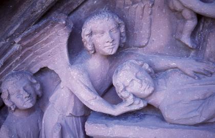 Aus Buntsandstein gefertigtes Tympanon über dem Westportal des Breisacher Münsters Nahaufnahme einer aus rötlich grauem Stein gehauenen Figurengruppe. Die mittlere von drei Figuren trägt Engelsflügel auf dem Rücken.