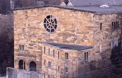 Ehemalige „Lausterhalle“ aus massivem Travertinmauerwerk Blick auf ein großes, aus rötlich braunen Mauersteinen gebautes Gebäude. In die Frontseite ist ein großes rundes Fenster eingelassen, das an ein Steuerrad erinnert. Davor ist ein kleineres Gebäude angebaut, aus dem gleichen Mauerwerk.