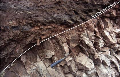 Erosionsdiskordanz in einer Baugrube in Friedenweiler im Eisenbach-Granit Blick auf flächiges, dunkles Gestein (links und oben) sowie bröckeliges, hellgraues Gestein (rechts sowie unten). Eine eingezeichnete schmale, diagonale Linie markiert die Grenze zwischen Grundgebirge und überlagernden Sedimentgesteinen.