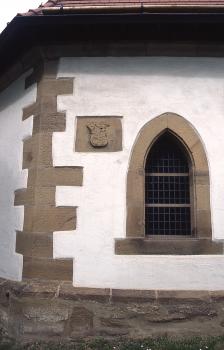 Friedhofskapelle in Güglingen aus Pfaffenhofener Sandstein Teilansicht eines kirchlichen Gebäudes mit Ecksteinen, Fensterumrandung und Wappen aus hellbraunem Mauerwerk. Sockel dunkelgrau, Wand weiß verputzt.