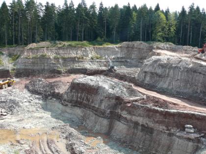 Gewinnung von Mürbsandstein in der Quarzsandgrube Birkhof Blick von erhöhtem Standort auf mehrere hintereinanderliegende, violettgraue Steinbruchwände. Die Brüche sind durch Fahrstraßen getrennt. Im Hintergrund stehen hohe Nadelbäume an.