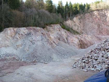 Durch eine Störung stark zerriebener Triberg-Granit am westlichen Rand des Steinbruch Tennenbronn (RG 7816-1) Steinbruchwand mit ocker- bis rotgrauem, absandendem Granit im linken Abschnitt und kompaktem rotgrauem Granit im rechten Abschnitt. Oberhalb der Steinbruchwand liegt ein Mischwald. Im Vordergrund rechts liegt ein Haufen mit Steinen aus dem Granitbruch.