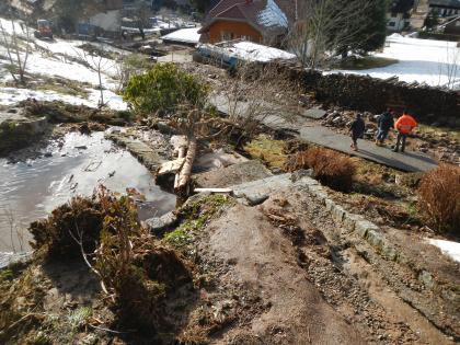 Abgelagerte Fracht aus Totholz, Geröll sowie einem Schluff/Sand-Gemisch in der bebauten Ortslage von Menzenschwand-Hinterdorf Blick von oben herab auf ein Grundstück, das von Schlamm, kleinen Steinen und Holzresten bedeckt ist. Dahinter ist eine Straße zu sehen, neben der größere Gesteinsbrocken liegen. Im Hintergrund stehen weitere Häuser.