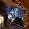 Blick in das Innere eines Besucherbergwerks. Rechts öffnet sich im Gestein ein höhlenartiger Raum, der bläulich angestrahlt und mit einem Gitter gesichert ist. Links unten führen Gleise in einen langen, ausgeleuchteten Schacht.