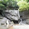 Blick auf steile graue Felswände, die links schräg aufeinanderliegen. Rechts fließt ein dünner Wasserfall zwischen den Felsen und bildet unten einen nach links führenden Bach mit steinigem Ufer.