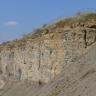Seitlicher Blick auf eine hohe, nach links hinten abfallende graugelbe Steinbruchwand. Die Kuppe ist mit Gras bewachsen und gesichert. Von rechts schiebt sich ein grauer Fließhang ins Bild.