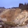 Seitlicher Blick auf die nach rechts geneigte Abbauwand einer Kiesgrube. Das braune Material weist im Vordergrund helle graue Stellen auf. Die Kuppe ist bewachsen. Links ist eine weitere Abbauwand zu sehen, sie hat eine graue Färbung.
