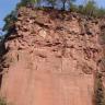 Blick auf eine hohe, rötlich braune Steinbruchwand. Die untere Hälfte der Gesteinswand ist glatt, in der oberen Hälfte türmen sich bankige Blöcke, die eine schartige Oberfläche bilden. Am oberen Ende der Wand wachsen Bäume.
