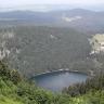 Blick von hoch oben über einen bewaldeten Steilhang auf einen tief unten liegenden, rechteckigen blauen See. Der See ist von Wald und angrenzenden bewaldeten Bergen umgeben. Links stechen Felsgrate durch den Wald, rechts ist eine freie Fläche mit Äckern.