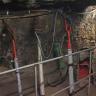 Das Bild zeigt mehrere historische Bohrmaschinen im Besucherbergwerk Teufelsgrund. Die Bohrgeräte sind mit Luftschläuchen verbunden und stehen hinter einem Schutzgitter an einer Stollenwand.