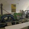 Blick in einen Ausstellungsraum des Besucherbergwerks Teufelsgrund mit heller, gewölbter Decke. Im Vordergrund steht eine große Fördermaschine mit blaugrauem Schwungrad.