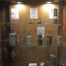 Blick auf eine hölzerne Vitrine mit Glasböden im Besucherbergwerk Teufelsgrund. Ausgestellt sind historische Grubenleuchten, wie etwa Karbidlampen sowie eine Froschlampe.