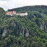 Blick auf einen bewaldeten Bergkegel. An den Seiten des Berges treten mehrere Felszacken hervor. Auf der flachen Kuppe des Berges stehen mehrere Gebäude, die miteinander verbunden sind.