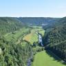 Blick aus großer Höhe über ein langgezogenes Tal mit Flusslauf. Das Tal ist hufeisenförmig von bewaldeten Bergen eingerahmt. Am Berghang links im Vordergrund stehen bleiche Felsen heraus.