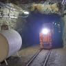 Das Bild zeigt eine beleuchtete Grubenbahn auf Schienen im Besucherbergwerk Finstergrund. Die Bahn - rechts im Bild - fährt durch einen schmalen, niedrigen Tunnel. Links an der Stollenwand ist ein Benzin- oder Öltank aufgestellt.