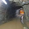 Blick in den tunnelartigen Durchgang eines Bergwerkstollens. Am Ende des Stollens, rechts der Bildmitte, ist ein beleuchteter Querstollen erkennbar.