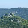 Blick auf einen bewaldeten Bergkegel mit einer Burgruine auf der Kuppe und vorstehenden Felsen etwas darunter. Im Hintergrund verlaufen bewaldete Berge mit ebenen Kuppen. An deren Hängen sind ebenfalls Felsformationen zu erkennen.