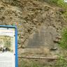 Hier sieht man eine steile rötlich braune, verwitterte Felswand, die von Bäumen und Sträuchern umgeben ist. Etwa in der Mitte der Felswand ist ein dunklerer, dreieckiger Bereich glatt geschliffen. Links ist noch ein Stück von einer Schautafel zu sehen.