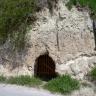 Das Bild zeigt eine hellgraue Lösswand vor einem Weganschnitt. Kuppe und linke Wandseite sind bewachsen. Am Fuß der Wand, unterhalb der Bildmitte, befindet sich eine vergitterte Öffnung.