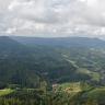 Blick aus großer Höhe auf bewaldete, gerundete Berge. Zwischen ihnen verteilen sich baumlose Flächen mit Siedlungen. Links erhebt sich ein höherer Berg mit abgeflachter Spitze. Rechts im Hintergrund sind weitere bewaldete Bergrücken erkennbar.