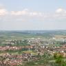 Blick aus großer Höhe über eine weite Landschaft mit einer Siedlung im Vordergrund. Zum Hintergrund hin sind Felder, Wiesen und bewaldete Hügel zu erkennen. Zwei dieser Hügel bilden ein Tal, das von einer langen, hohen Brücke überspannt wird.