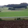 Panoramabild einer weiten, gewellten Landschaft mit Äckern und Grünflächen. Die Kuppen sind bewaldet. Im Vordergrund ist ein dunkelbrauner, erhöht liegender Ackerstreifen.