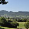 Blick über eine hügelige Landschaft mit Bäumen, Wiesen, Wald und fernen Siedlungen. Im Hintergrund erheben sich von rechts flache, bewaldete Höhen mit scharfer Kante. Weiter links und mittig liegen eine kleine und dann eine größere runde Bergkuppe auf.