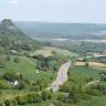 Von erhöhtem Standort blickt man auf eine wellige Landschaft mit Wiesen, Äckern, Bäumen und Wald. Links, hinter einer bewaldeten Anhöhe, erhebt sich ein ebenfalls bewaldeter Bergkegel mit Burgruine. Rechts führt eine kurvige Autobahn am Berg vorbei.